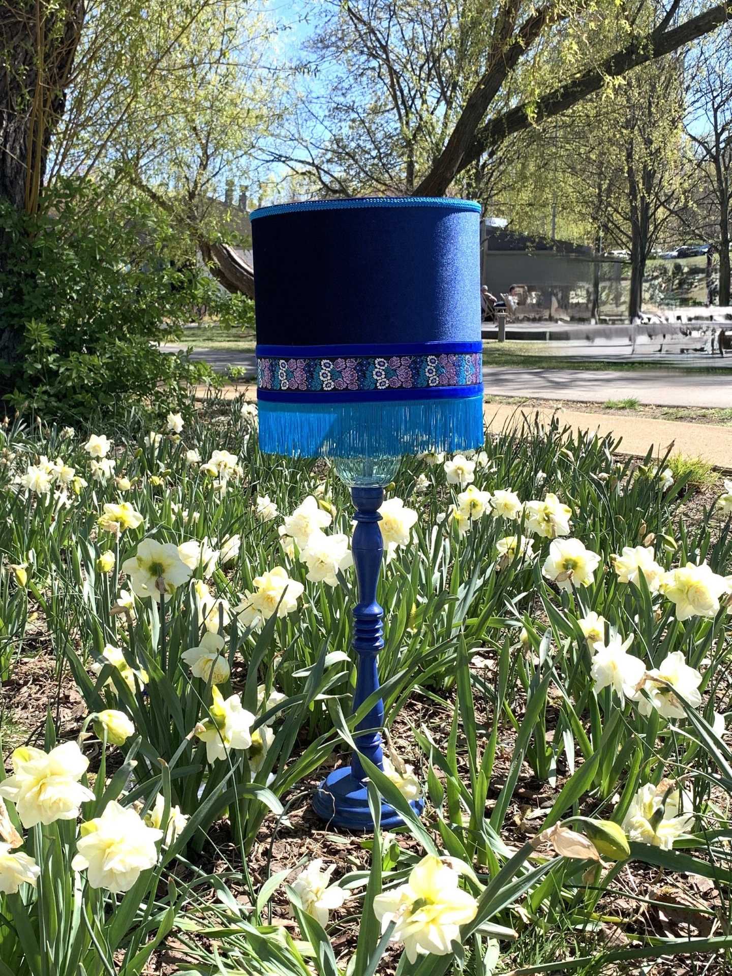 Lampa stojąca granatowa z granatowym abażurem. Klosz ma frędzle i dekoracyjną pasmanterię w kwiatki.