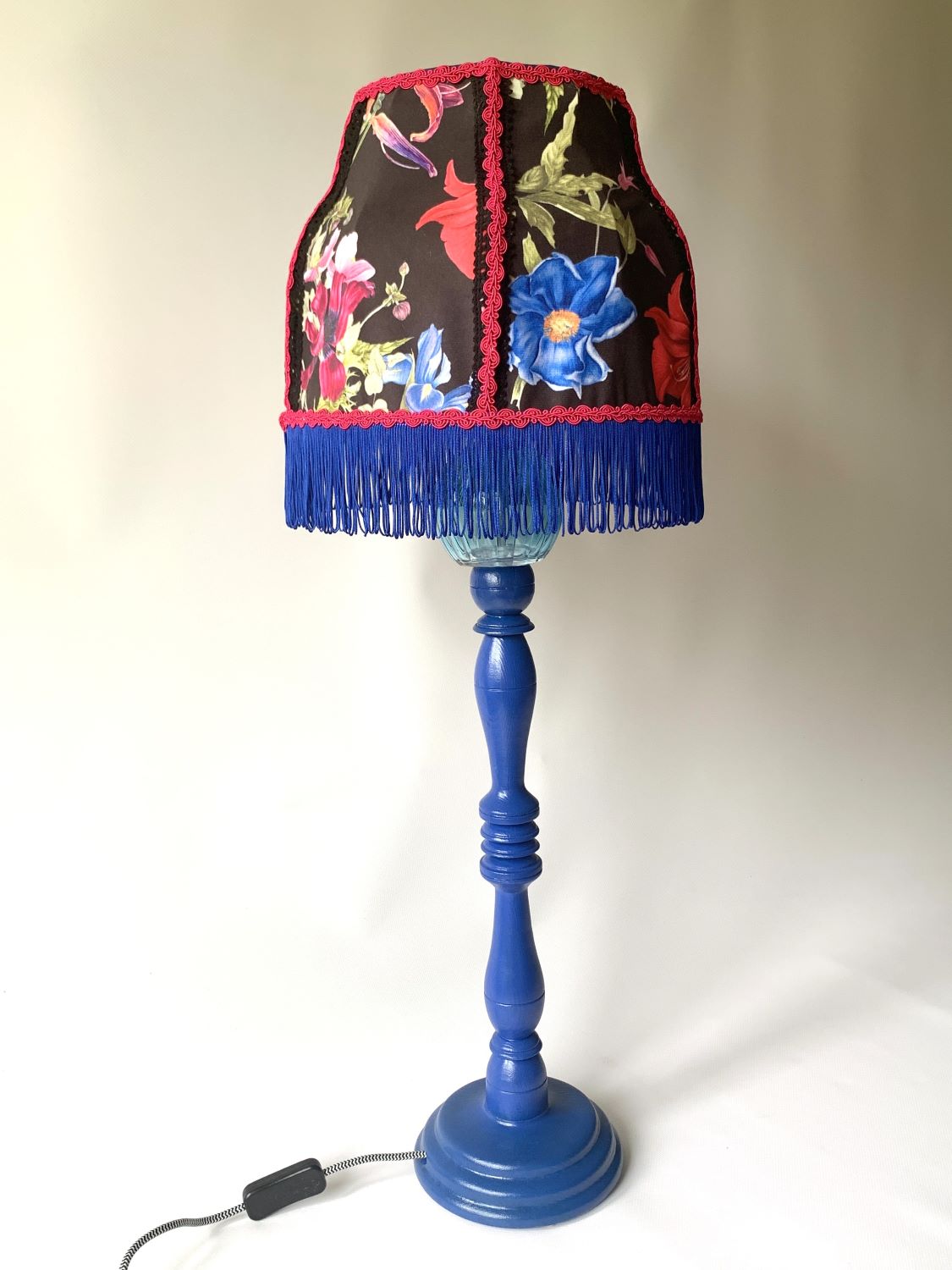 Stojąca granatowa lampa z abażurem w kwiaty na czarnym tle i dekorowanym frędzlami.