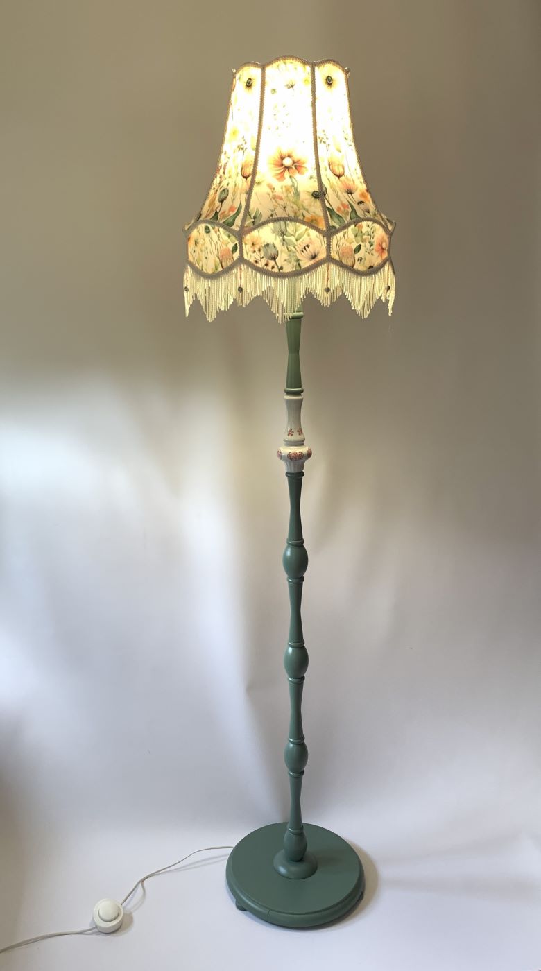 Wysoka lampa podłogowa z zieloną podstawą i abażurem w kwiaty z zapaloną żarówką.